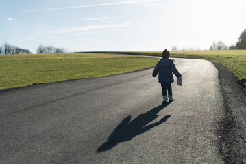 Obraz premium Beautiful winter photo of a little warmly dressed girl with a shadow on a rural asphalt winter road without snow, rear view