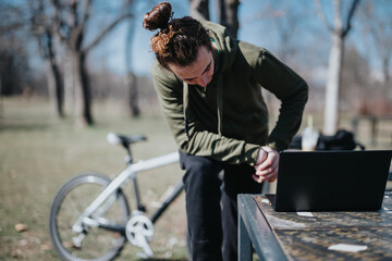 An active male takes a break at a park table to check his smart watch while his bicycle rests...