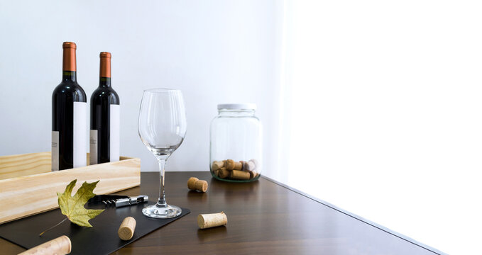 Copa de cristal vac&iacute;a con botellas de vino tinto, sacacorchos y corchos dispersos sobre la mesa. 