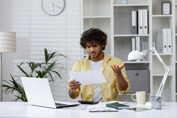 Confused man looking at a document in a modern office, expressing frustration. Surroundings include...