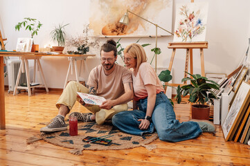 Artistic painter couple sitting on atelier floor and looking at artwork.