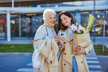 A smiling young woman helps her elderly mother with shopping bags, sharing a smartphone screen...