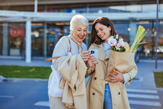 A smiling adult woman guides her cheerful senior mother through smartphone use after grocery shopping, both dressed in casual attire, united in a moment of learning and bonding.