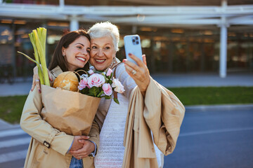 A joyful adult daughter and her beaming senior mother engage with a smartphone outside a store, a...