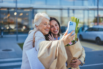 A joyful senior mother and her adult daughter pose for a selfie, phone in hand, outside a grocery...