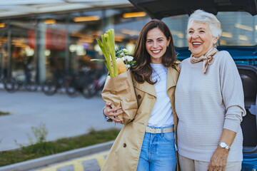 A smiling young woman and her cheerful elderly mother stand with fresh groceries outside a store,...