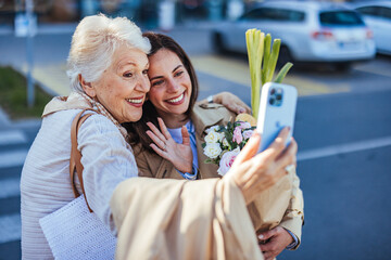 A young adult female helps her elderly mother, as they share a joyful moment taking a selfie with a...