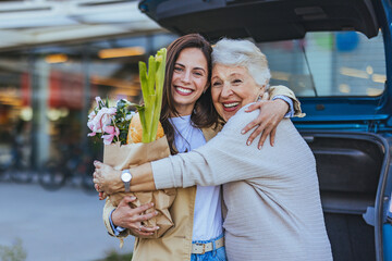 A joyful adult daughter embraces her elderly mother, holding a bag of fresh groceries beside a car,...