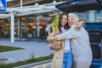 A smiling adult woman embraces her joyful elderly mother, loaded with fresh groceries, displaying...