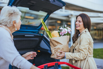 Caucasian adult daughter loads groceries into car trunk, smiling as she assists her elderly mother,...