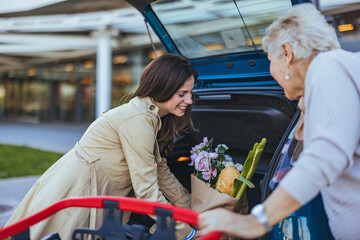 A Caucasian adult daughter is cheerfully loading groceries into the car trunk, assisting her...