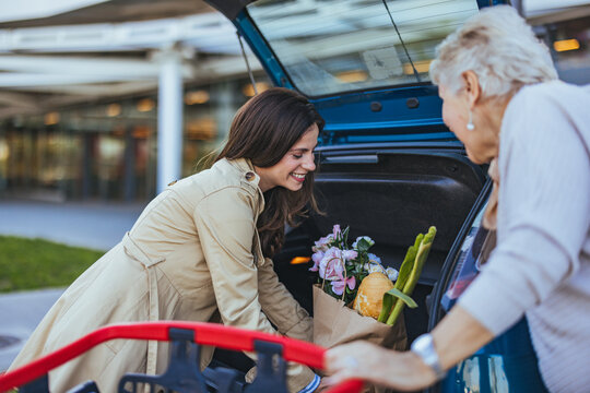 A Caucasian adult daughter is cheerfully loading groceries into the car trunk, assisting her elderly mother outside a shopping center.