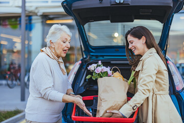 A cheerful Caucasian woman helps her elderly mother with grocery bags, placing them into the car...