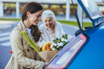 A smiling Caucasian woman is placing a bag filled with groceries into a car trunk, assisted by her...