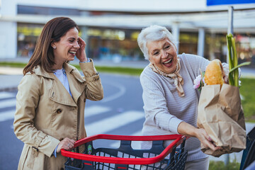 A smiling Caucasian woman assists her joyful senior mother, placing fresh groceries into the car...