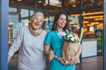 A smiling Caucasian nurse aids an elderly woman carrying a grocery bag, embodying support and...
