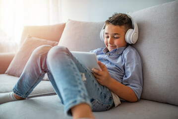 Smiling boy with tablet PC and headphones listening to music or playing game at home. Happy Little...