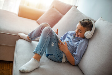 The 6 year old boy is wearing white headphones and is using tablet. Sunny day. Stock photo of a...