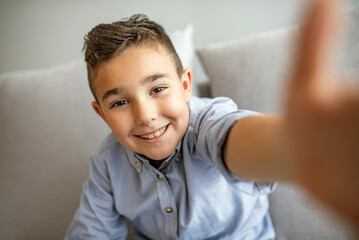 Cute little boy taking selfie on light background. Happy little boy with healthy teeth taking selfie. Photo of boy takes selfie with smart phone indoors. Boy takes selfie