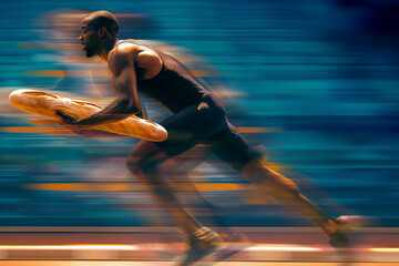 A dark-skinned man running holds a large French loaf in one hand. The atmosphere is competitive, at a sporting event