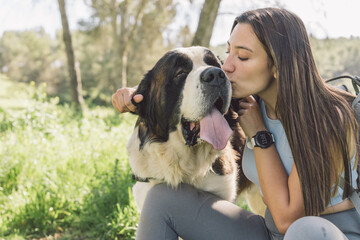 Woman kissing happy dog in nature. Joyful woman affectionately kisses her saint bernard dog at a...