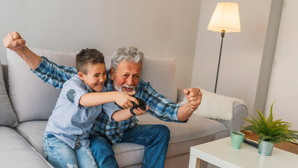 Grandfather and grandson are playing video games at home. Young boy is playing games with elder...