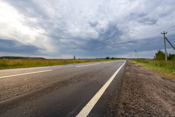 A long, straight asphalt road stretches through a rural landscape under a dramatic, overcast sky.