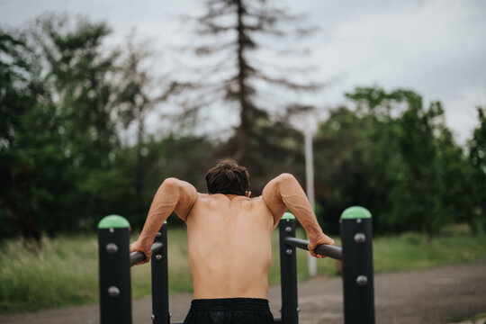 Back view of a man doing an outdoor calisthenics workout on parallel bars in a park. Focus on strength and fitness.