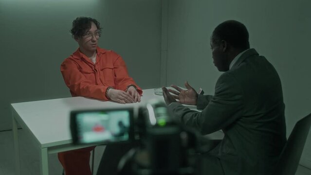 Medium full shot of African American male detective questioning detained Caucasian violent crime suspect at police station, asking questions, video camera recording interrogation in blurred foreground