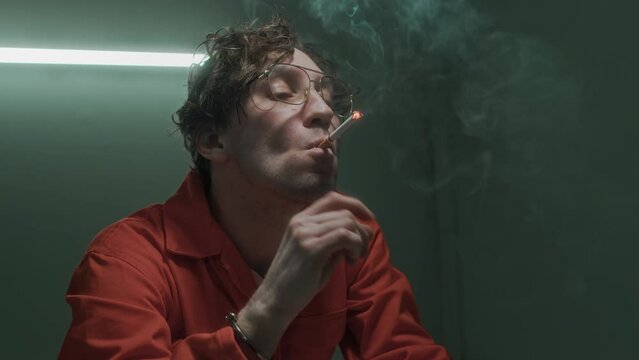 Medium close-up shot of Caucasian male murder suspect asking police officer to light his cigarette, then drawing, letting out puff of smoke, and taunting detective, while being interrogated in prison