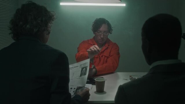 Medium rear shot of diverse police detectives preparing to interrogate man in handcuffs, smoking cigarette, detained for violent crime, female agent looking at document with profile photo
