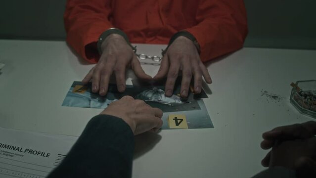 Medium close-up shot of hands of unrecognizable federal agent laying out crime scene photographs in front of detained suspect in handcuffs, prison suit, who is studying them with interest and talking