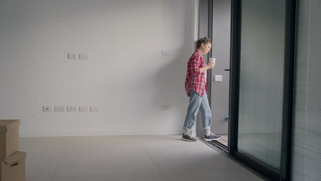 Female holding coffee cup looking outside in empty apartment or new office. Excited and smiling with her new home or moving out. Small cardboard boxes on the floor. Empty clean walls all around