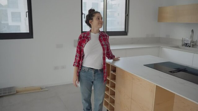 Woman looking around her new apartment then leans on kitchen island satisfied. Female home owner impressed with the new design and potential of brand new house or home feeling happy observing it