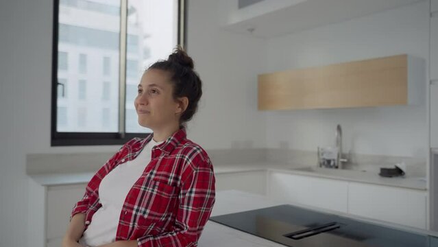 Happy young woman leaning on kitchen island in her brand new apartment. After moving into new home looking around satisfied with her place. Cardboard boxes on the table as she visualize the potential