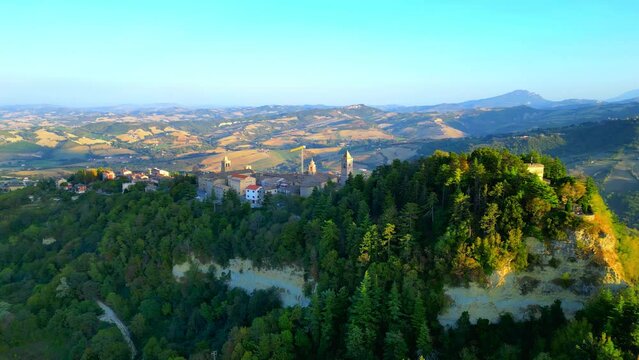 Sumptuous aerial truck left and pan right shot from Penna San Giovanni (MC), an overview of the town with its majestic vegetation-filled cliff soaring above the surrounding typical Marche hillscapes