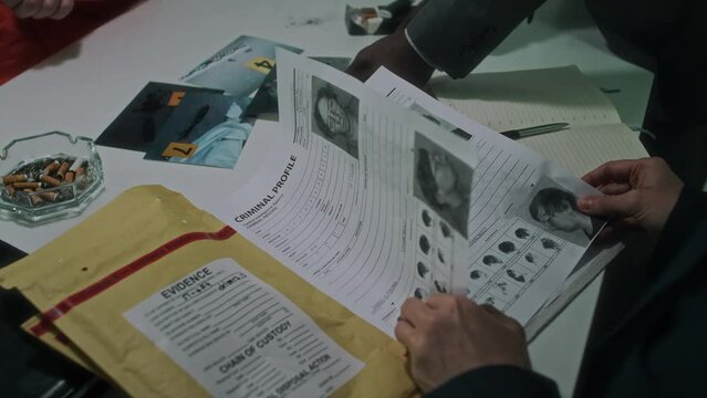 Close-up over-shoulder shot of hands of unrecognizable diverse federal agents looking at criminal profiles, demonstrating photos of dead body to anonymous suspect in handcuffs, smoking cigarette