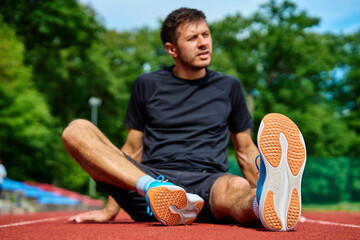 Male athlete wearing running shoes sits on stadium track, resting and catching his breath after run...
