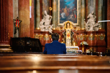 Back view of man in blue jacket prays in catholic church. He is seated in pew with his head bowed...