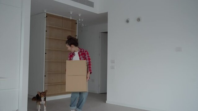 Young woman walking into her new apartment with her small dog carrying boxes. Happy and excited entering empty home for the first time. Dancing with her dog in their brand new place