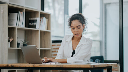 Professional Young Woman Working on Laptop in Modern Office Setting with Bookshelves and Large...