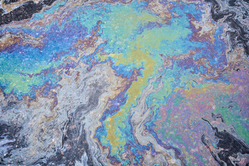 A rainbow-hued slick of spilled engine oil and gasoline spreads across the asphalt surface,...