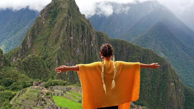 Girl in orange woven poncho raises arms taking in views of Machu picchu