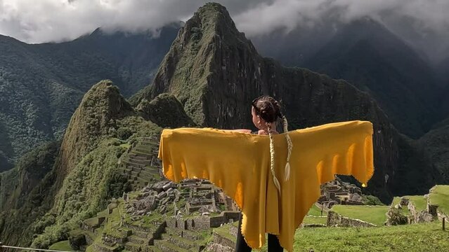Woman raises arms wearing yellow poncho in front of Machu picchu, rearview