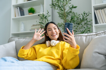 Happy young asian woman  wearing headset and relaxing on comfortable couch at home, texting...