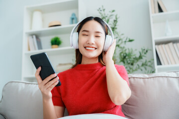 Young Asian woman relaxing on sofa at home, listening music with headset on cell phone. People...