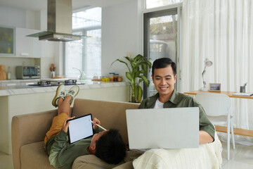 Smiling man working on laptop from home when son drawing on tablet screen