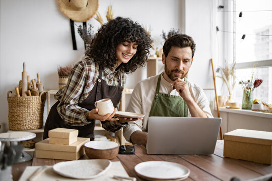 Caucasian couple of co-workers work on small pottery production business. Mature man and young curly-haired woman in aprons use laptop to take inventory of products on store website.