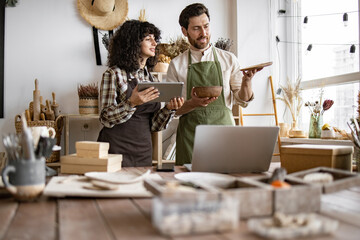 Two Caucasian workers in uniform doing inventory together at decor shop. Bearded mature man counting goods while young curly woman using tablet.