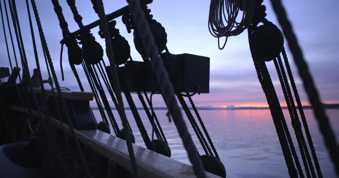 1700s sailing ship underway at sunrise. Slow pan across ratlines. Water and sunrise in soft focus.
